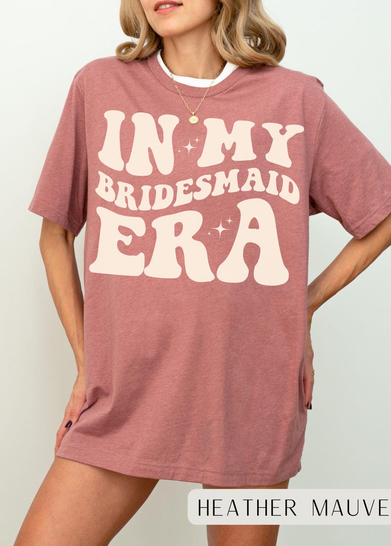 May include: A heather mauve t-shirt with the text "In My Bridesmaid Era" in a retro font with sparkling stars.