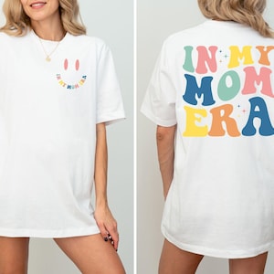 Puede incluir: Camiseta blanca de gran tamaño con un gráfico de cara sonriente en la parte delantera y el texto "In My Mom Era" en letras retro de colores en la parte posterior.