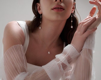 以下が含まれることがあります： 白いドレスを着て、透き通る袖、真珠のネックレスとイヤリングをしている女性。彼女は椅子に座って、手を前に組んでいます。