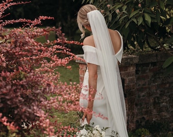 May include: A bride in a white off-the-shoulder gown with a long veil, holding a bouquet of white flowers. She is standing in a garden with lush greenery and red foliage.