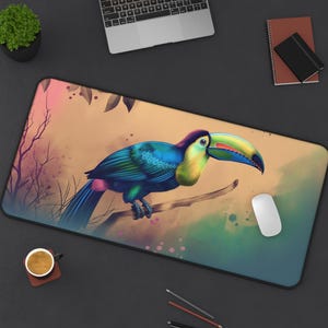 Toucan desk mat with tropical watercolor bird artwork on a large rectangular mouse pad. A colorful toucan with a large beak sits on a branch in a jungle setting. The toucan is blue, green, yellow, and orange. The background is a watercolor wash of pink, green, and brown.
