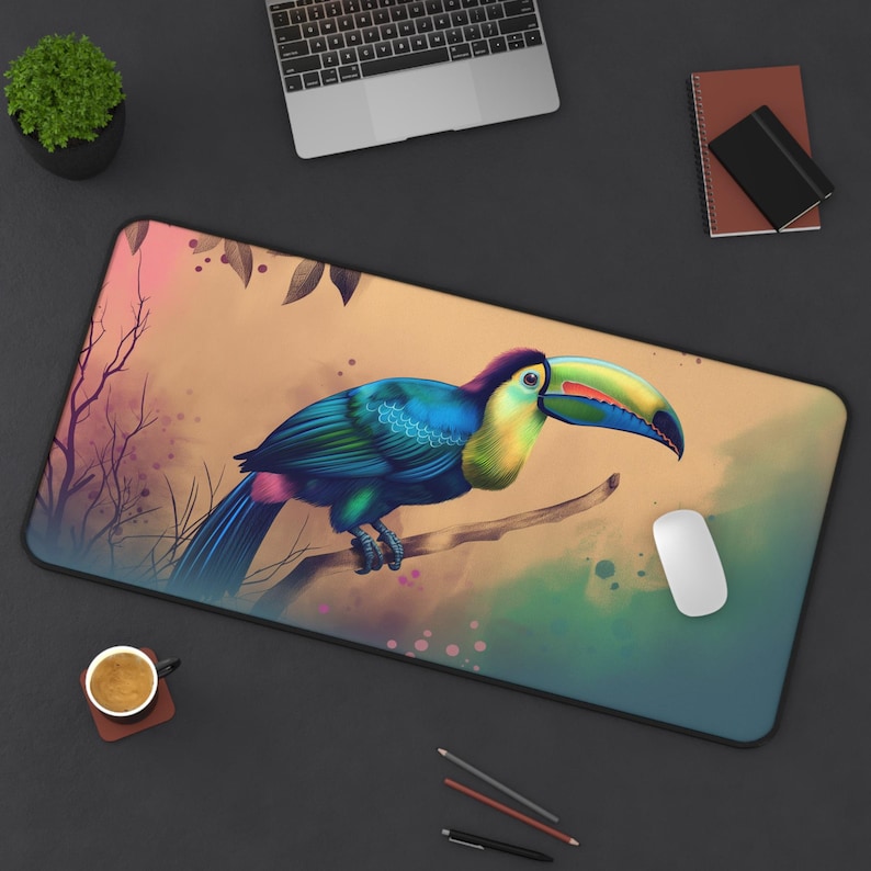 Toucan desk mat with tropical watercolor bird artwork on a large rectangular mouse pad. A colorful toucan with a large beak sits on a branch in a jungle setting. The toucan is blue, green, yellow, and orange. The background is a watercolor wash of pink, green, and brown.