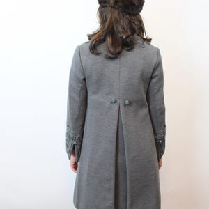 May include: A gray coat with decorative sleeve cuffs and button details. The coat has a tailored fit and a pleated back. A brown fur hat is worn on the head.