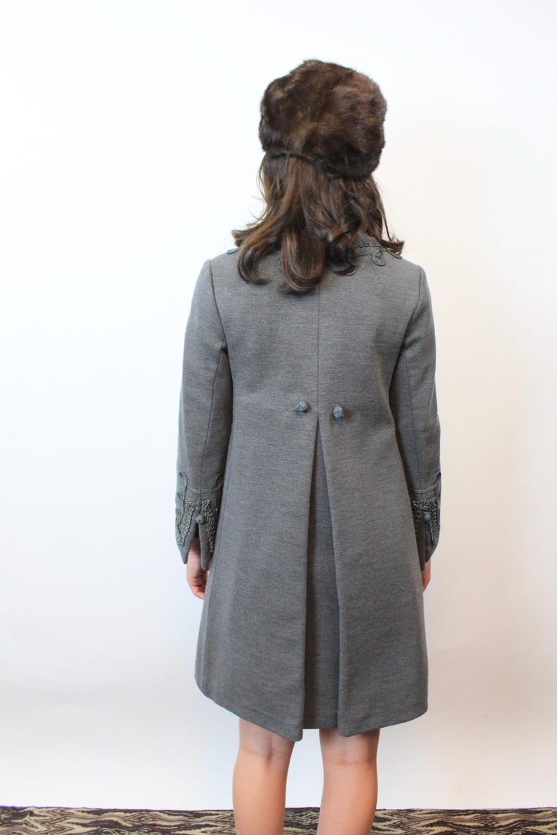 May include: A gray coat with decorative sleeve cuffs and button details. The coat has a tailored fit and a pleated back. A brown fur hat is worn on the head.