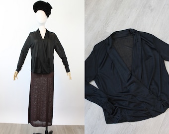 May include: A black long-sleeved wrap top with a V-neck design. The top is displayed on a mannequin with a black hat and a brown skirt. The fabric appears to be lightweight and flowing. The second image shows the top on a wooden surface.
