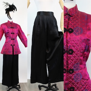 May include: A magenta quilted jacket with black frog closures and a mandarin collar, paired with black wide-leg pants. The jacket features a pattern of blue and purple designs. A black feathered headpiece is also visible.