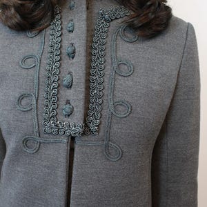 May include: A gray jacket with decorative teal trim and buttons. The jacket features a high collar and ornate teal rope detailing around the neckline and front closure. The design includes looped accents.