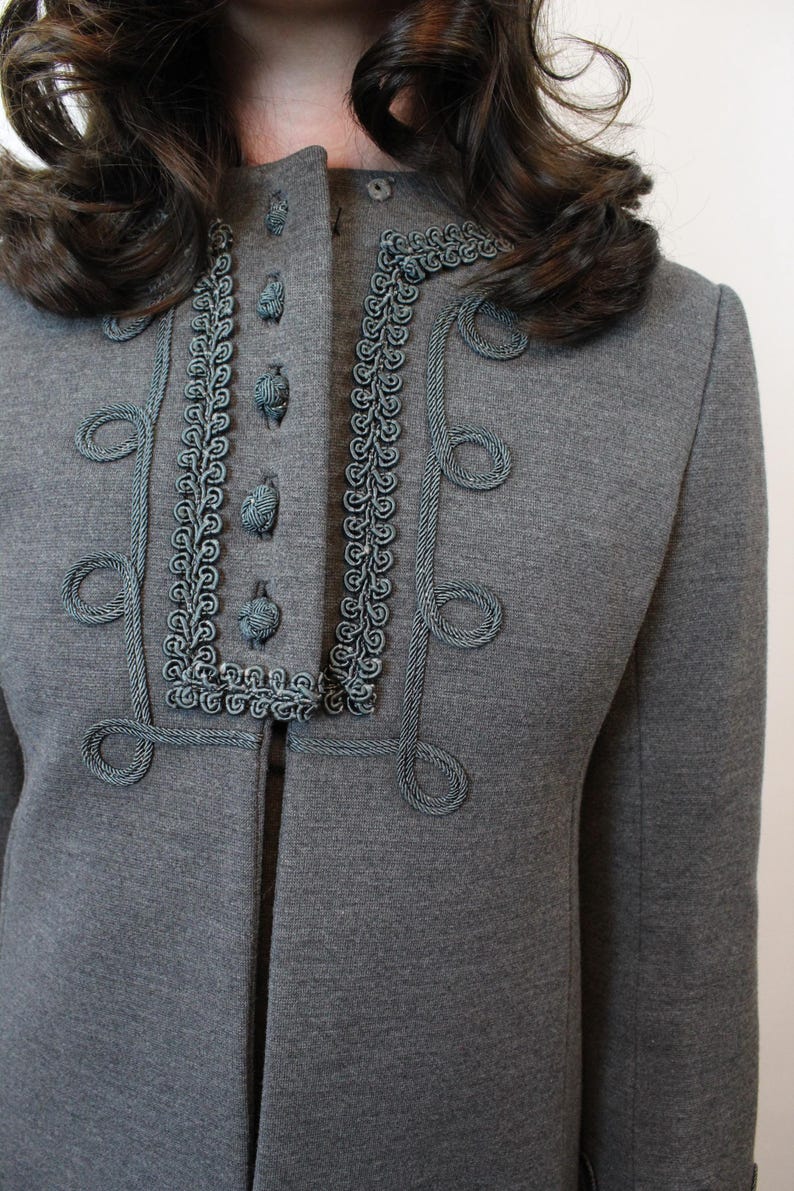 May include: A gray jacket with decorative teal trim and buttons. The jacket features a high collar and ornate teal rope detailing around the neckline and front closure. The design includes looped accents.