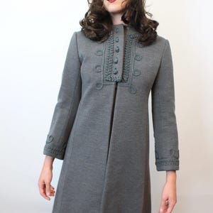 May include: A gray vintage coat with decorative trim around the collar, cuffs, and button details. The coat has a flared silhouette and long sleeves. The model is wearing a brown fur hat. The coat is a classic piece of outerwear.