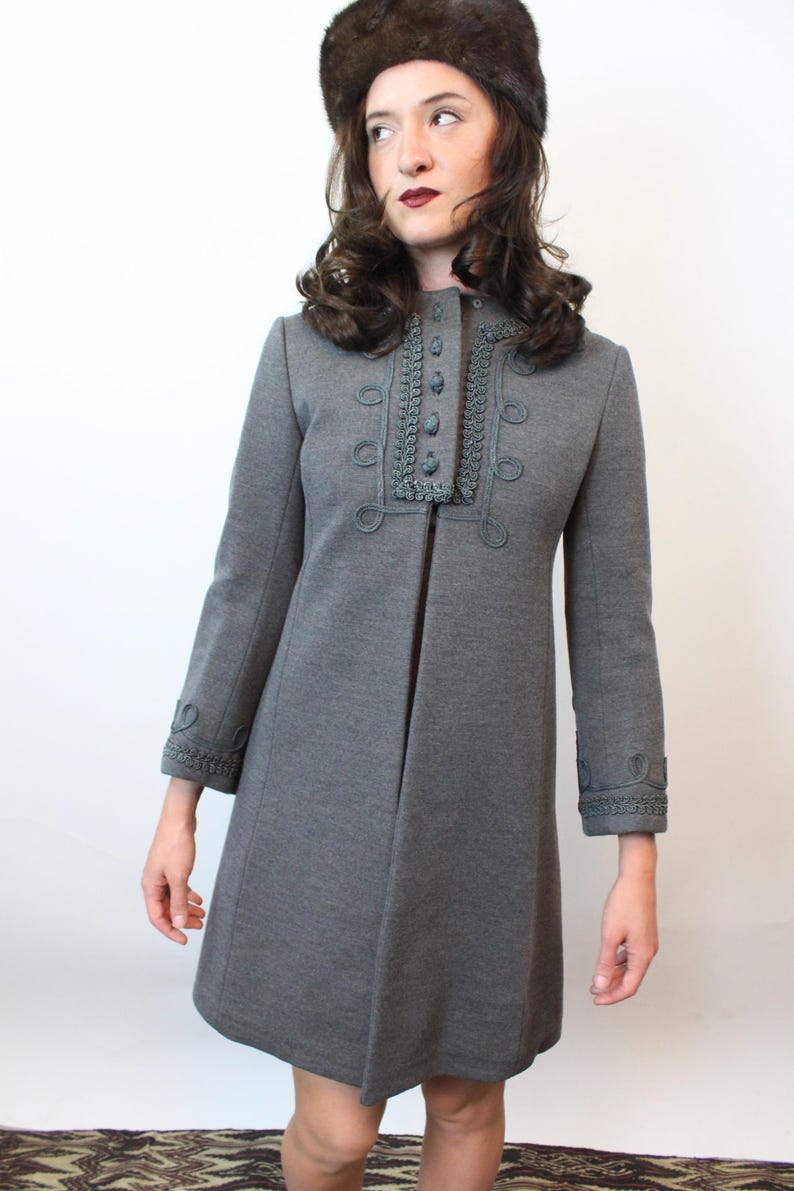 May include: A gray vintage coat with decorative trim around the collar, cuffs, and button details. The coat has a flared silhouette and long sleeves. The model is wearing a brown fur hat. The coat is a classic piece of outerwear.