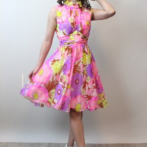 May include: A pink, yellow, and purple floral print dress with a high neck and a fitted bodice. The dress has a flared skirt and is made of a lightweight fabric. The model is wearing white shoes.