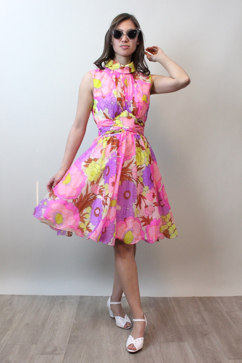 May include: A pink, yellow, and purple floral print dress with a high neck and a fitted bodice. The dress has a flared skirt and is made of a lightweight fabric. The model is wearing white shoes.
