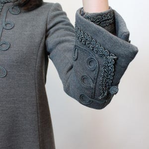 May include: Gray coat with decorative trim. The coat features a high collar and ornate detailing around the cuffs and front. The cuffs are folded back, revealing a contrasting lining. The coat has a classic, tailored design.