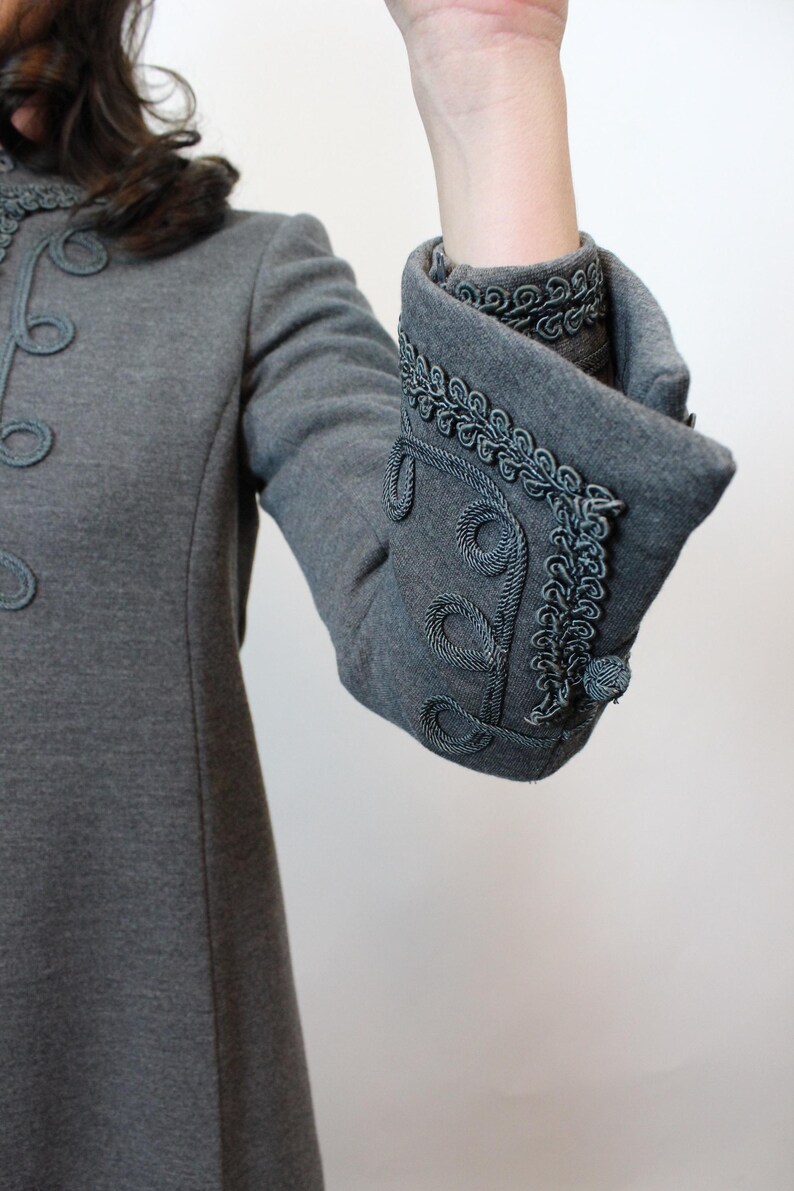 May include: Gray coat with decorative trim. The coat features a high collar and ornate detailing around the cuffs and front. The cuffs are folded back, revealing a contrasting lining. The coat has a classic, tailored design.