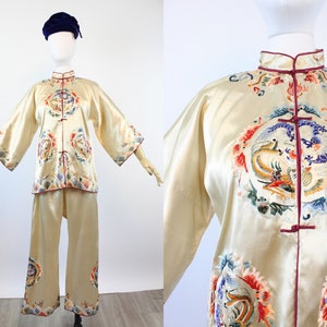 May include: Cream-colored silk pajama set with intricate embroidery. The jacket and pants feature colorful dragon and phoenix designs, with red trim. The set includes a mandarin collar jacket and wide-leg pants, displayed on a mannequin.