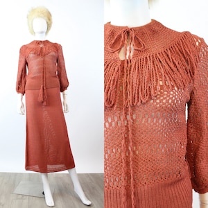 May include: A rust-colored knitted two-piece outfit, consisting of a long-sleeved top with a fringed collar and a matching midi skirt. The top features a tie closure and a tassel. The outfit is displayed on a mannequin.