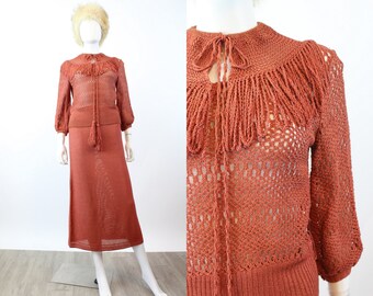 May include: A rust-colored knitted two-piece outfit, consisting of a long-sleeved top with a fringed collar and a matching midi skirt. The top features a tie closure and a tassel. The outfit is displayed on a mannequin.