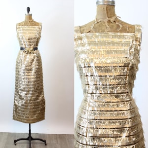 May include: A vintage gold-colored sleeveless dress with horizontal fringe detailing. The dress is displayed on a mannequin and cinched at the waist with a black belt. The dress has a floor-length hemline.