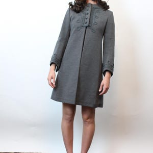 May include: A gray, knee-length coat with decorative stitching around the collar and cuffs. The coat has a row of buttons down the front and is paired with gold-colored shoes. The person is wearing a brown fur hat.