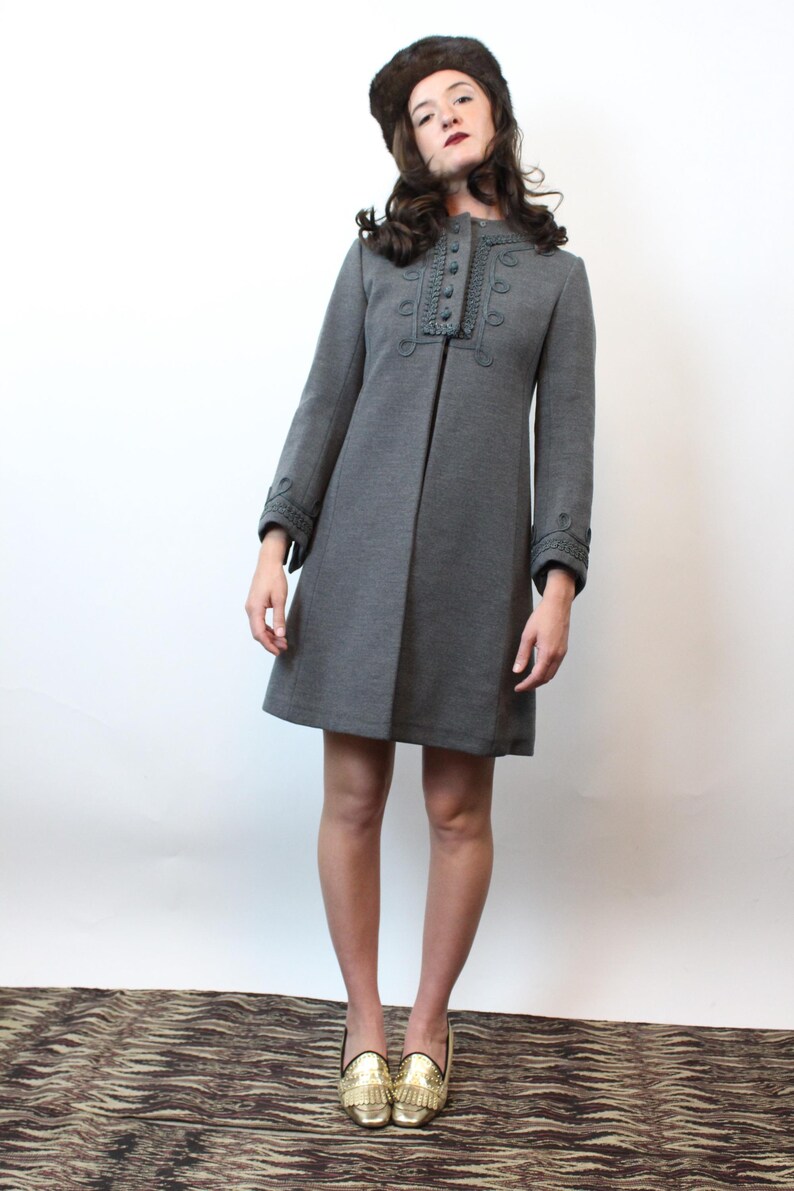 May include: A gray, knee-length coat with decorative stitching around the collar and cuffs. The coat has a row of buttons down the front and is paired with gold-colored shoes. The person is wearing a brown fur hat.