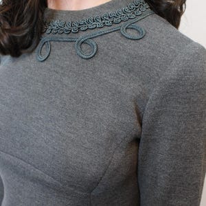 May include: A gray dress with a high neckline and long sleeves. The neckline is adorned with a decorative trim in a matching gray color, featuring a rope-like design with circular accents. The dress has a tailored fit.