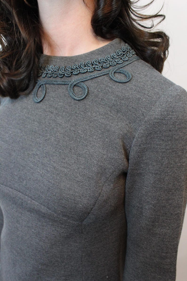 May include: A gray dress with a high neckline and long sleeves. The neckline is adorned with a decorative trim in a matching gray color, featuring a rope-like design with circular accents. The dress has a tailored fit.