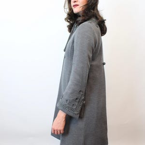 May include: A gray coat with decorative trim on the cuffs and collar. The coat has a button closure and long sleeves. The person in the image is also wearing a brown fur hat. The coat appears to be made of a soft, woven material.