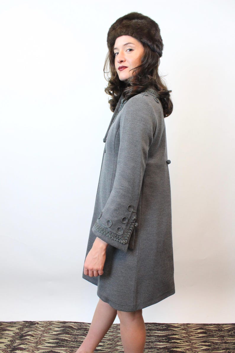 May include: A gray coat with decorative trim on the cuffs and collar. The coat has a button closure and long sleeves. The person in the image is also wearing a brown fur hat. The coat appears to be made of a soft, woven material.