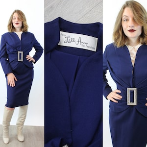 May include: A navy blue skirt suit with a matching jacket and knee-length skirt. The jacket features a decorative rhinestone buckle at the waist. The label inside the jacket reads "Lilli Ann Paris San Francisco".