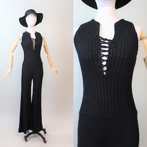 Puede incluir: Mono negro de punto con cuello halter y detalle de cordones. El mono tiene perneras anchas y se exhibe en un maniquí con un sombrero negro. La tela parece ser un punto acanalado.