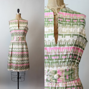 Puede incluir: Un vestido sin mangas con un patrón geométrico en tonos de verde, rosa y dorado. El vestido tiene un escote en V y un cinturón en la cintura.