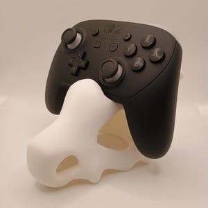 Op de afbeelding: Een zwarte Nintendo Switch Pro-controller rustend op een witte, driedimensionale schedelvormige standaard. De controller is voorzien van standaardknoppen, twee analoge sticks en het Nintendo Switch-logo in het midden van de matzwarte behuizing.