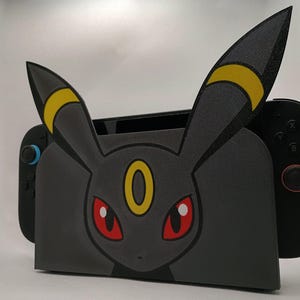 May include: A black gaming console dock featuring a stylized Umbreon character design with large pointed ears, yellow ring markings, and red eyes. The dock is shaped to fit a handheld gaming device with black controllers attached on both sides.