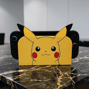 May include: A yellow Pikachu-themed gaming console dock featuring the character's face with black-tipped ears, red cheeks, and a small smile. The dock is black with a blue button on the left controller and a red button on the right controller.