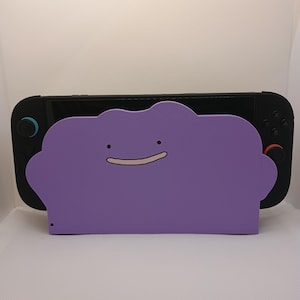 May include: A purple, cloud-shaped console dock cover featuring a simple, smiling face with two small black dots for eyes and a thin, curved line for a mouth. The cover is positioned over a black gaming console with black and red controllers.
