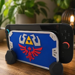 May include: A black gaming console held in a blue, silver, and red shield-shaped dock. The dock features a silver border, a central yellow triangle emblem, and a red bird-like crest. The console is supported by two black, faceted rock-shaped stands.
