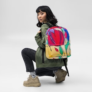 Puede incluir: Una mochila colorida con un vibrante diseño floral en tonos rojos, morados y amarillos. La mochila tiene la espalda y las correas negras. El patrón floral incluye hojas y tallos verdes. La mochila la lleva una persona con una chaqueta verde y vaqueros negros.