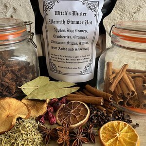 May include: A collection of aromatic ingredients for a simmer pot, including dried orange slices, cinnamon sticks, star anise, and cloves. A bag labeled "Witch's Winter Warmth Simmer Pot" is in the background, along with two glass jars.