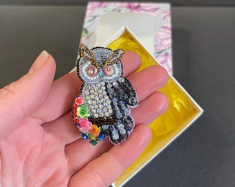 May include: A grey owl brooch with black and white feathers, sparkling rhinestones, and colourful flowers. The owl has large, round eyes with red pupils.