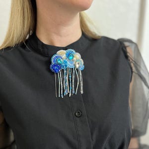 May include: A beaded brooch in shades of blue, white, and gold, with a cloud-like design and dangling beaded fringe. The brooch is attached to a black button-down shirt.