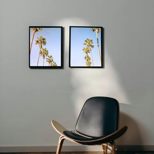 Op de afbeelding: Twee ingelijste zwart-witfoto's van palmbomen tegen een blauwe lucht, hangend aan een witte muur boven een zwarte leren stoel met houten poten.