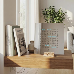 Könnte beinhalten: Ein modernes Holzregal zeigt Bücher, eine Vase und eine einzigartige Uhr. Das Zifferblatt der Uhr hat ein Raster aus Buchstaben mit hervorgehobenen Wörtern. Zu den Büchern gehören "BAUHAUS" und "ART & DESIGN". Ein kleiner Holzblock mit dem Wort "HOLZ" ist ebenfalls vorhanden.