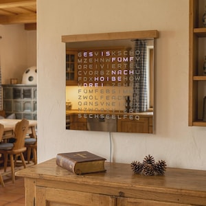 Könnte beinhalten: Eine moderne Digitaluhr mit Holzrahmen zeigt die Zeit mit leuchtenden Buchstaben an. Die Uhr ist an einer cremefarbenen Wand über einem Holzschrank angebracht. Ein geschlossenes Buch und Tannenzapfen liegen auf dem Schrank.