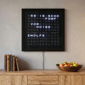 Könnte beinhalten: Eine schwarze, quadratische Wortuhr zeigt die Uhrzeit in weißen, leuchtenden Buchstaben an. Die Uhr ist an einer hellen Wand über einem Holzschrank angebracht. Eine Holzschale mit Obst und ein Stapel Bücher befinden sich auf dem Schrank.