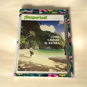 May include: A magazine titled "¡Despertad!" with the headline "Cómo Calmar El Estrés" is encased in a clear plastic sleeve. The sleeve is bordered with a green fabric featuring a floral pattern. The magazine cover depicts a beach scene.