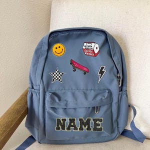 May include: A blue backpack with a variety of colourful patches, including a smiley face, a skateboard, a lightning bolt, and a star. The backpack has a large pocket on the front with the word "NAME" in black letters.