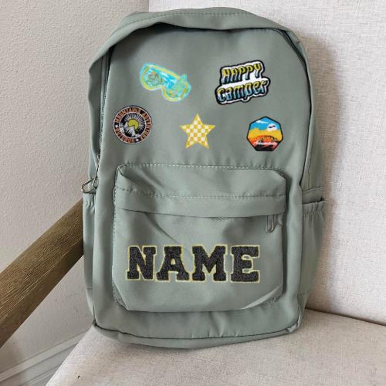 以下が含まれることがあります： カラフルなパッチがついた緑色のバックパック。パッチには「Happy Camper」、星、太陽などがあります。バックパックの前方には大きなポケットがあり、黒い文字で「NAME」と書いてあります。