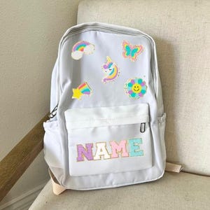 Puede incluir: Una mochila gris con una cremallera blanca y un parche que dice "NAME" en letras de arcoíris. La mochila tiene varios parches coloridos, incluyendo un arcoíris, una mariposa, un unicornio, una estrella y una flor.