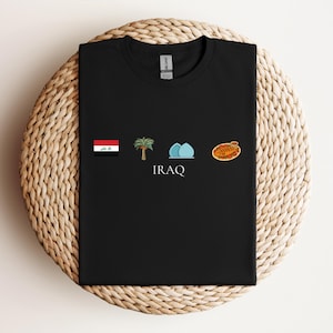 Può includere: T-shirt nera con un design che incorpora la bandiera irachena, una palma, due cupole blu e un piatto tradizionale. La parola "IRAQ" è stampata sotto le illustrazioni. La maglietta è esposta su una superficie circolare intrecciata.