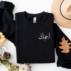 May include: Black crewneck sweatshirt with white Arabic script on the chest. The sweatshirt is displayed on a white surface with fall-themed decorations, including pumpkins, leaves, and a hat.